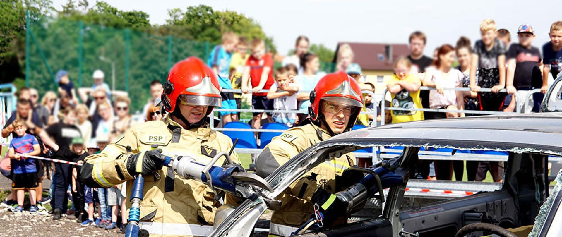 Zdjęcie przedstawia strażaków którzy tną sprzętem hydraulicznym wrak pojazdu w czasie pokazu dla dzieci.