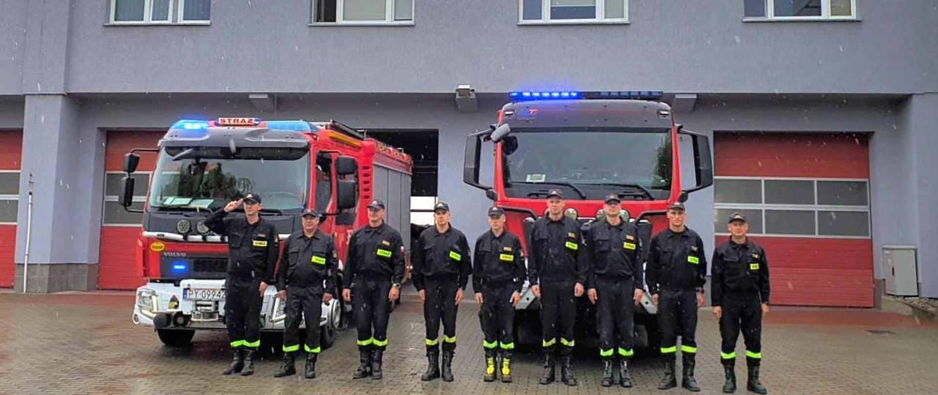 Zdjęcie przedstawia funkcjonariuszy oddających hołd bohaterom, stojącym na tle samochodów pożarniczych w włączonymi sygnałami świetlnymi i dźwiękowymi. Samochody stoją przez garażami z czerwonymi bramami.