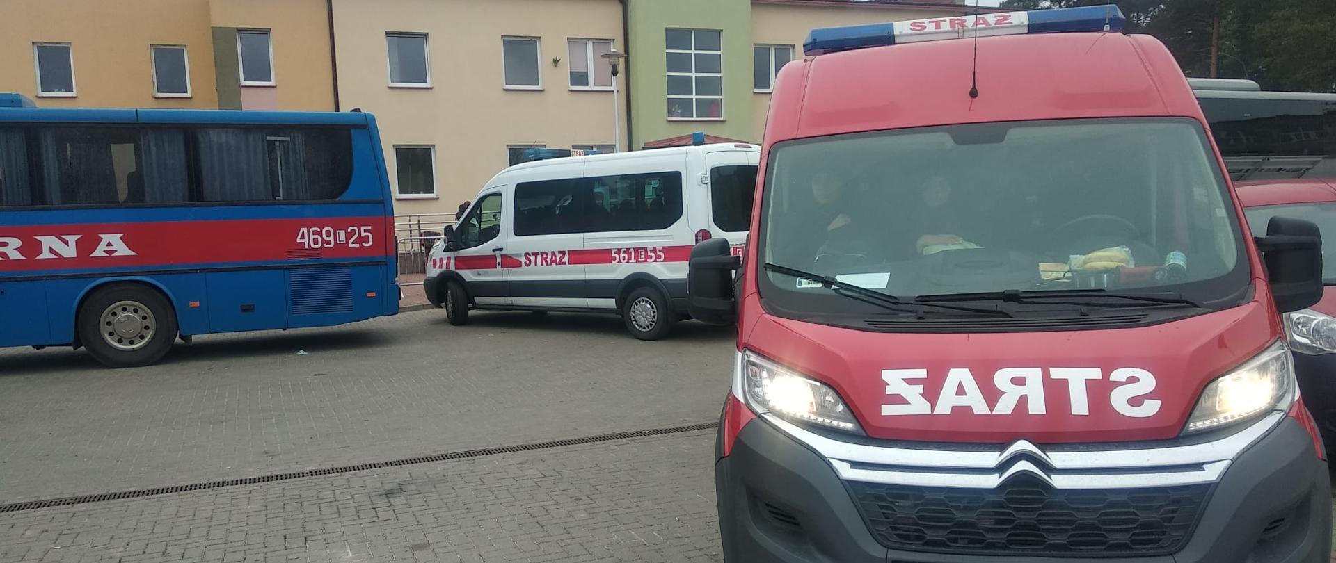 Zdjęcie przedstawia dwa busy straży pożarnej, jeden biały drugi czerwony do przewozu osób na granicy, obok stoi autokar koloru niebieskiego