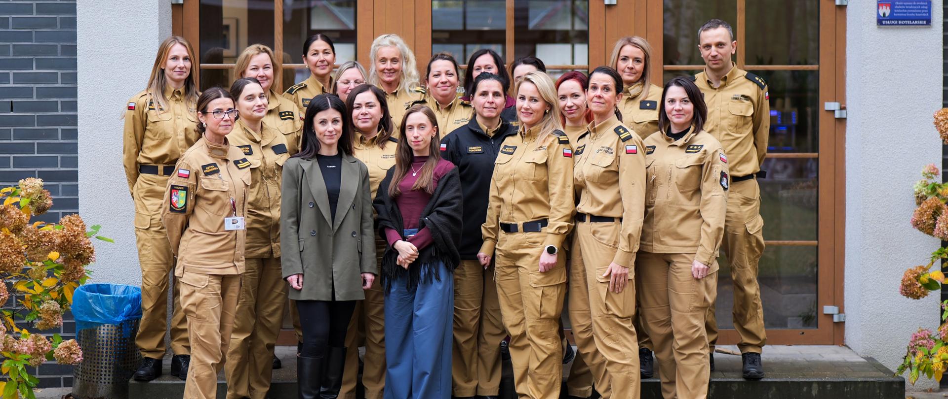 Grupowe zdjęcie uczestników odprawy kadrowej PSP woj. lubuskiego. Na pierwszym planie funkcjonariuszki i pracownicy cywilni Państwowej Straży Pożarnej w mundurach służbowych stoją przed budynkiem ośrodka szkoleniowego w Dankowie.