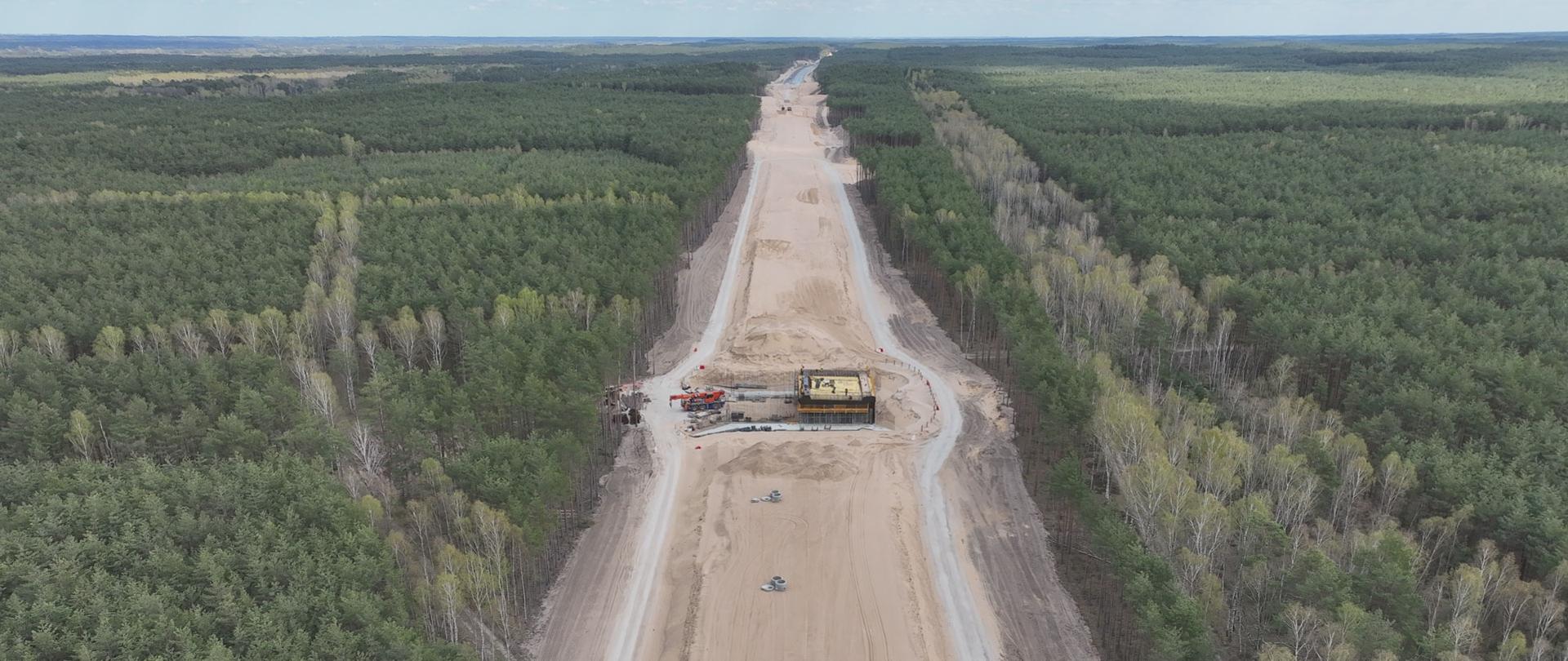 Widok z lotu ptaka na szeroki pas budowy drogi przebiegający przez gęsty las. W centrum kadru znajduje się prosty odcinek drogi w trakcie realizacji, pokryty jasnym piaskiem, z widocznymi śladami maszyn budowlanych oraz pojedynczymi betonowymi elementami ułożonymi na powierzchni. W połowie długości widoczny jest plac robót z konstrukcją mostową lub przepustem oraz sprzętem budowlanym, w tym dźwigiem. Po obu stronach przyszłej jezdni znajdują się tymczasowe drogi techniczne. Otaczający teren to rozległy, zielony las z przewagą drzew iglastych oraz pasami jaśniejszych drzew liściastych. Horyzont rozciąga się w oddali pod lekko zachmurzonym niebem. 