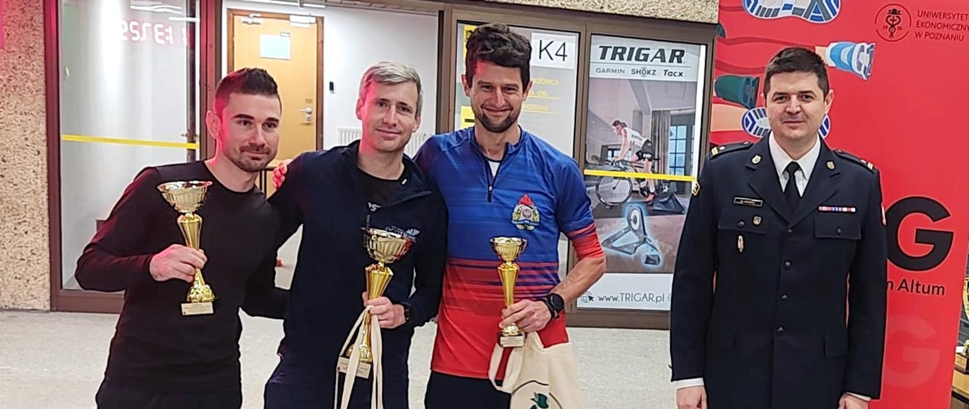 trzech mężczyzn w ubraniach sportowych stoi obok siebie z pucharami w ręku, obok stoi strażak w mundurze wyjściowym , w tle różne banery