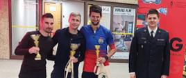 trzech mężczyzn w ubraniach sportowych stoi obok siebie z pucharami w ręku, obok stoi strażak w mundurze wyjściowym , w tle różne banery