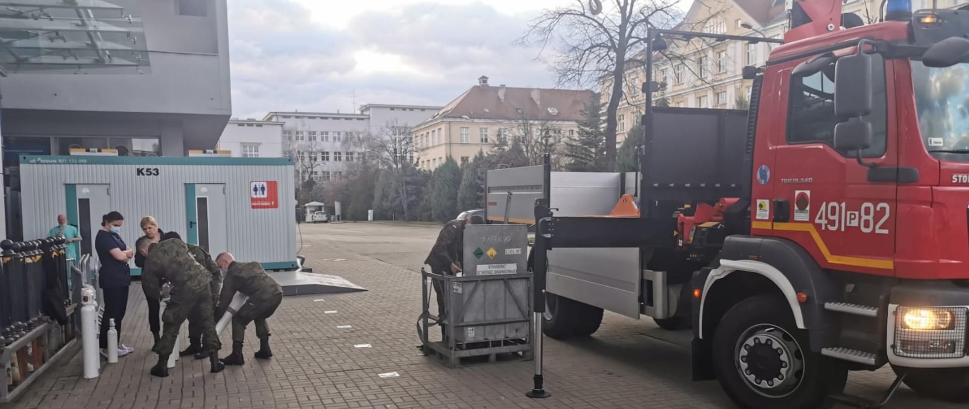 Wóz strażacki rozładowuje butle z tlenem obok żołnierze przenoszą butle do budynku.