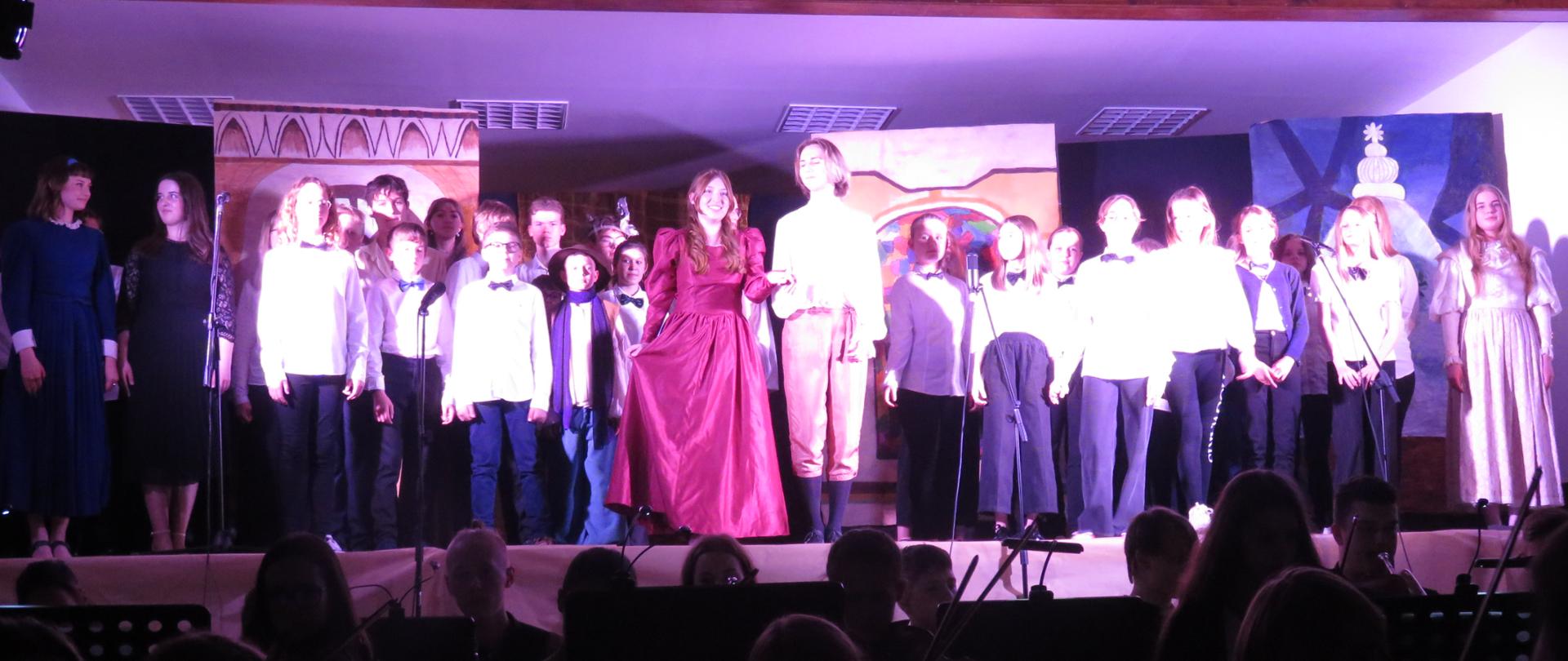 Wykonawcy stoją na scenie, orkiestra siedzi poniżej. 