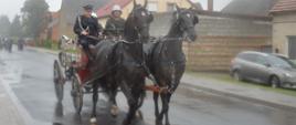 Jubileusz 95-lecia OSP Konary. Droga przez miejscowość, którą podąża sikawka. Ciągną ją dwa konie. Na sikawce znajduje się trzech strażaków. Za sikawką w oddali widać maszerującą kolumnę strażaków. W tle zabudowania i drzewa.