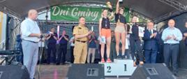 Zdjęcie przedstawia biegaczy podczas wręczenia nagród. Trzy kobiety stoją na podium i trzymają w ręce puchary. Obok stroją władze Gminy Rzgów oraz mundurowi. 