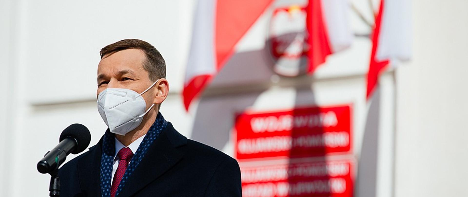 Premier Mateusz Morawiecki przemawia podczas uroczystości w tle budynek i flagi.