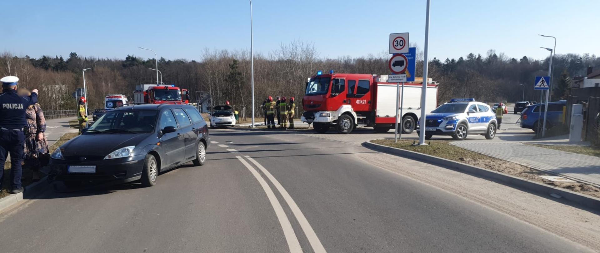 Rozbity samochód uczestniczący w wypadku stoi na ulicy. W tle samochody służb - policja, straż karetka - uczestniczące w działaniach.