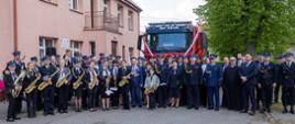 Zdjęcie grupowe uczestników uroczystości. W pierwszym szeregu stoi Marszałek Województwa Mazowieckiego, Zastępca Mazowieckiego Komendanta wojewódzkiego PSP st. Piotr Gąska i członkowie orkiestry dętej z instrumentami. W tle widoczny jest fragment samochodu strażackiego i część budynku. Wszyscy stoją na placu przed budynkiem. Za nimi rośnie wysokie drzewo z zielonymi liśćmi. 
