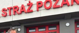 Dwaj uśmiechnięci strażacy w piaskowych mundurach pozują przed budynkiem straży pożarnej. Jeden z nich trzyma „Certyfikat Wsparcia”, a drugi stoi obok bordowego roweru i unosi kciuk w górę. W tle widać kolarzy.