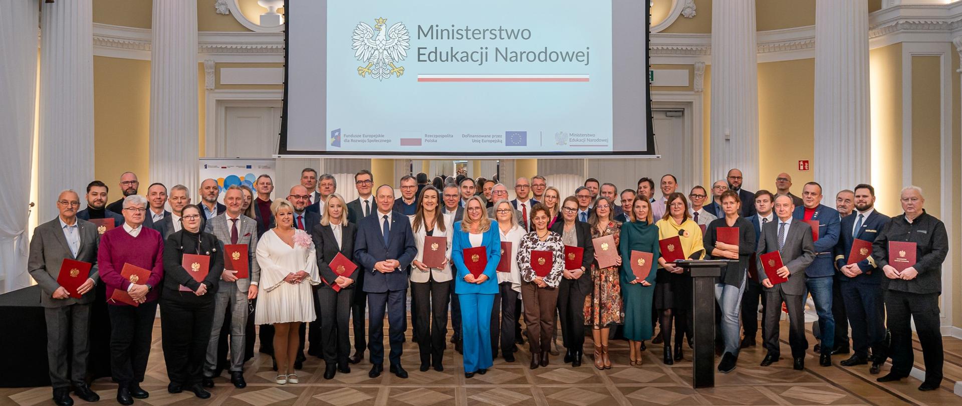 Duże grupowe zdjęcie uczestników wydarzenia, w tym wiceministra Henryka Kiepury, stojących na tle ekranu z napisem "Ministerstwo Edukacji Narodowej" i godłem Polski. Wszyscy trzymają czerwone teczki.
