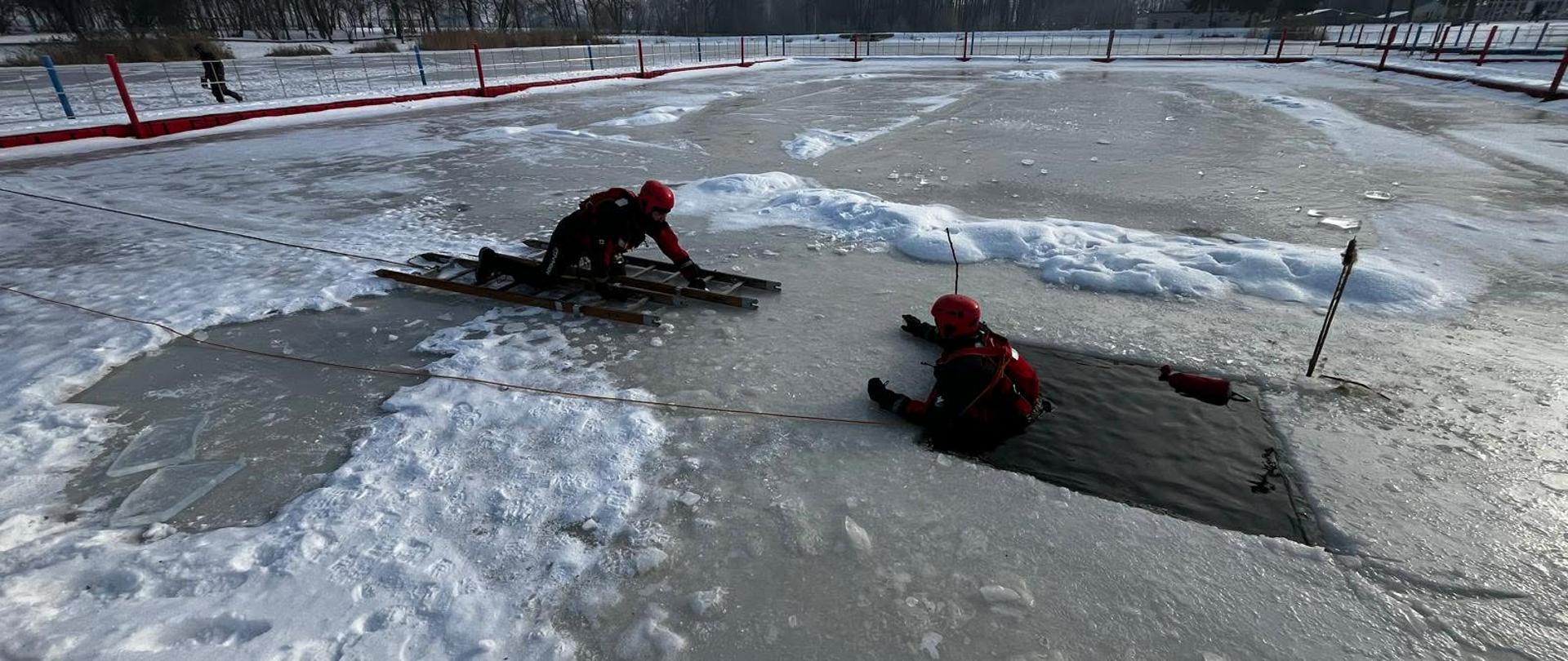 Ćwiczenia ratownictwa lodowego na zamarzniętym zbiorniku wodnym. Dwóch ratowników w czerwonych kaskach i skafandrach pracuje przy przeręblu — jeden znajduje się w wodzie, drugi leży na lodzie na drewnianych podporach, trzymając linę asekuracyjną. W tle rozległa, zaśnieżona tafla lodu oraz ogrodzenie i zimowy krajobraz z drzewami.