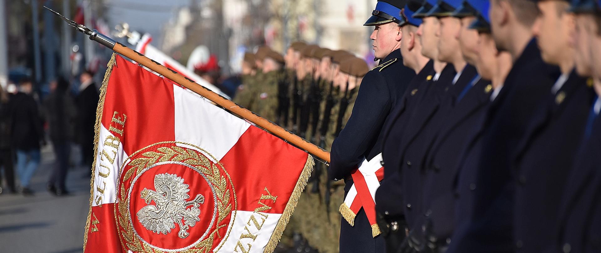 na zdjęciu poddział strażaków w granatowych płaszczach ustawiony bokiem do fotografującego, jeden ze strażaków trzyma opuszczony sztandar Komendy Wojewódzkiej PSP w Łodzi, w tle pododdział wojska i kilka osób, budynki, tło rozmazane