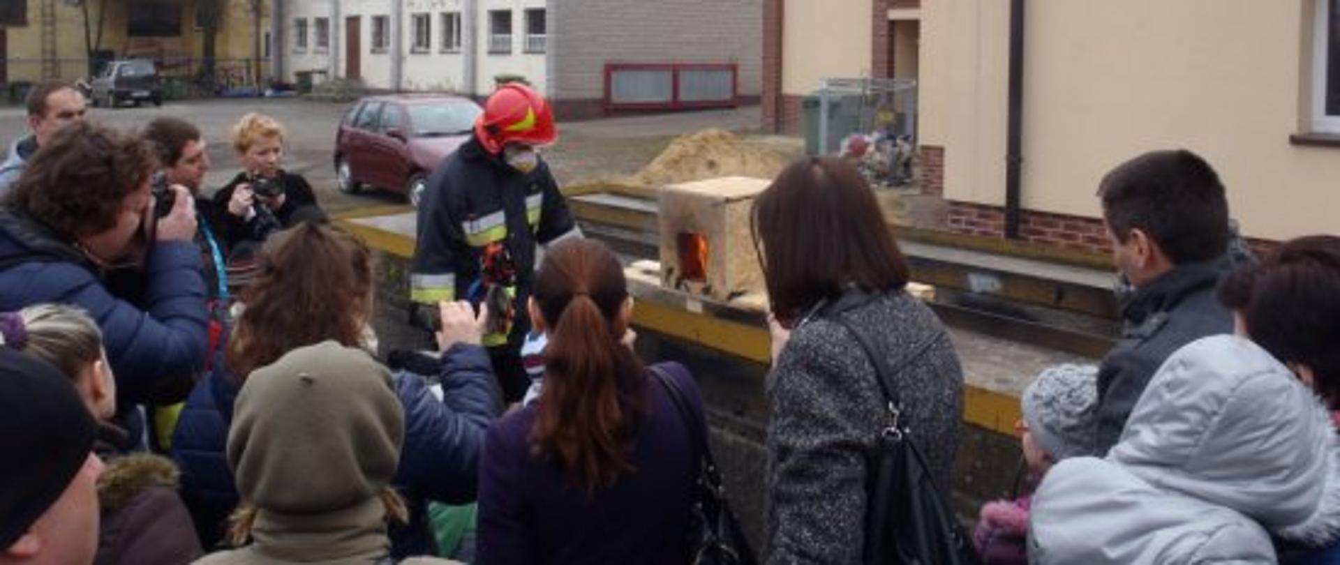 Zdjęcie ukazuje stanowisko symulacji pożaru w budynkach w postaci makiety domku ogniowego. Na pierwszym planie grupa kilkunastu osób obserwuje rozwój pożaru w makiecie przed nimi. Niektórzy z obserwatorów robią zdjęcia aparatami fotograficznymi i telefonami komórkowymi. Obok modelu stoi strażak ubrany w ubranie specjalne i maseczkę filtrującą.
