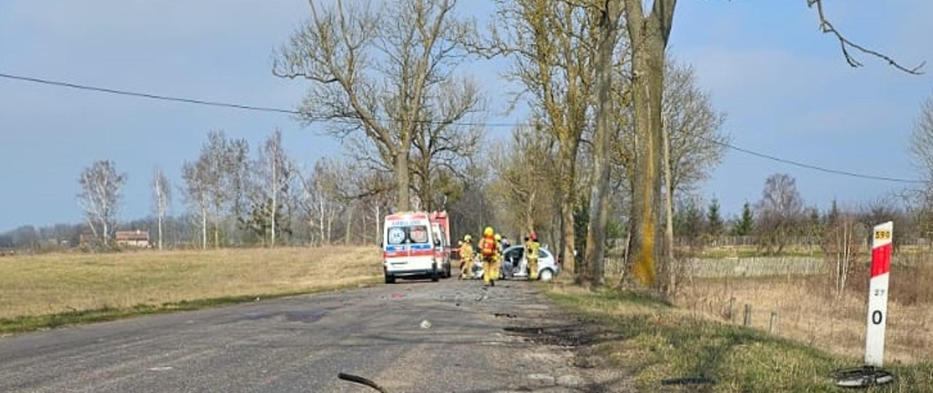 Na pierwszym planie znajdujące się na drodze urwane od samochodu koło wraz z zawieszeniem. W tle miejsce akcji ratowniczej – karetka, pojazd straży pożarnej oraz rozbity samochód osobowy, wokół nich strażacy. 
