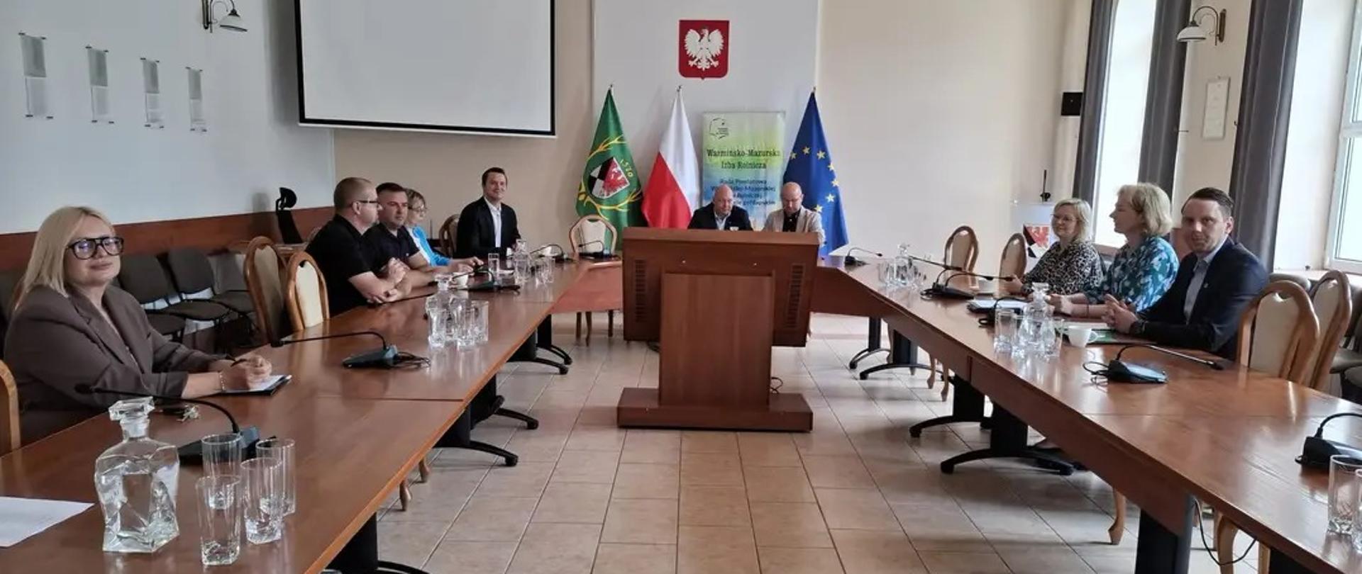 Grupa osób uczestniczących w spotkaniu siedzących przy stole. W tle baner Warmińsko-Mazurskiej Izby Rolniczej.