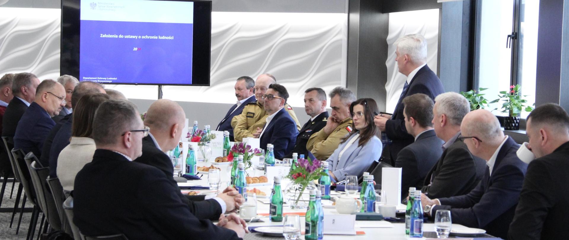 Uczestnicy odprawy zasiadający za stołem wysłuchują prezentacji założeń do ustawy, W tle na telewizorze wyświetlana jest prezentacja