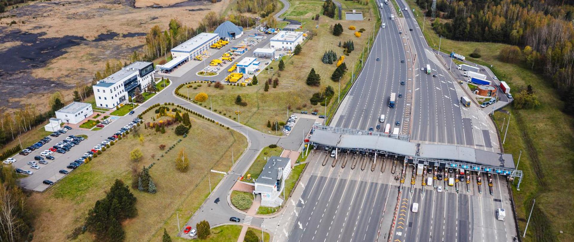Lotnicze ujęcie Punktu Poboru Opłat Brzęczkowice na autostradzie A4. Widoczna rozbudowana infrastruktura bramek z wieloma pasami ruchu oraz oznakowaniem poziomym kierującym ruch pojazdów. Po lewej stronie znajdują się budynki zaplecza technicznego i parking z zaparkowanymi samochodami, a wokół tereny zielone i przemysłowe.