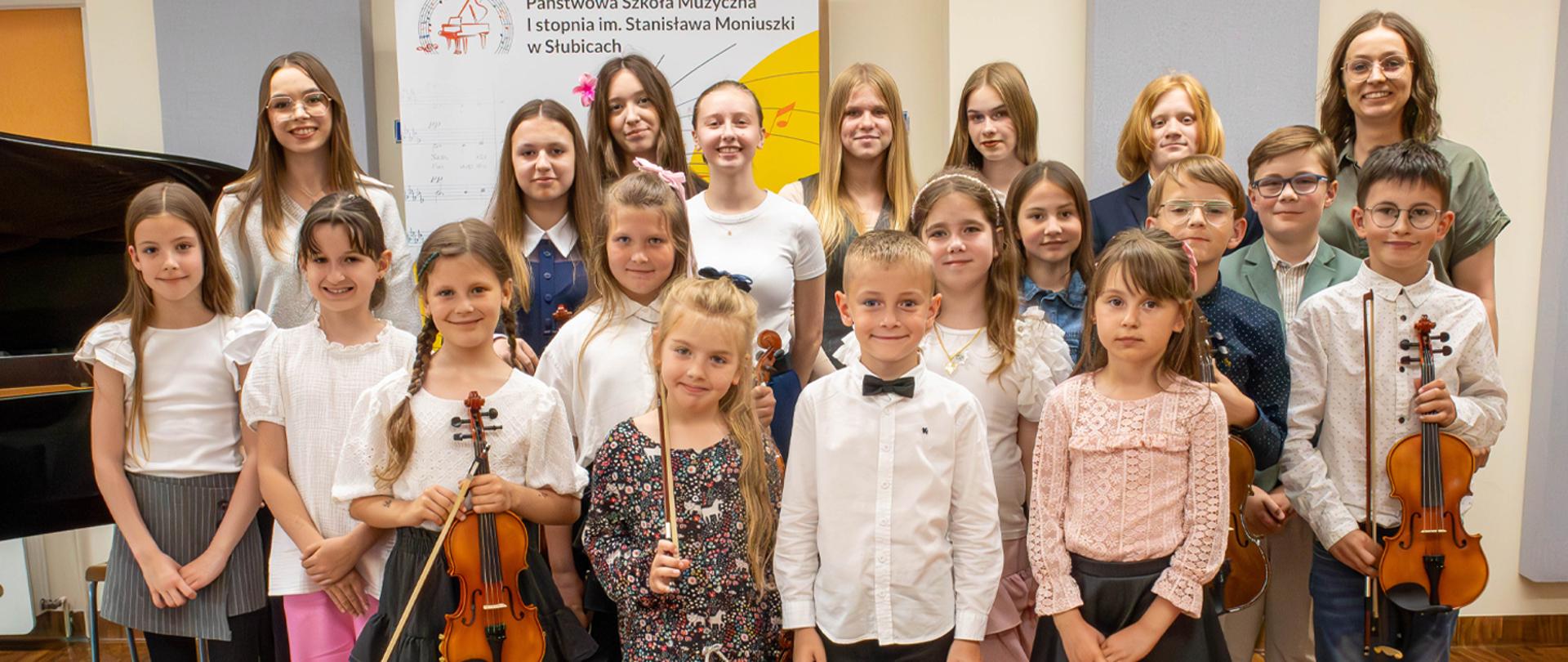 Grupa kilkunastu uczniów wraz z dwoma nauczycielkami stoją w auli szkoły na tle baneru szkolnego. Niektóre dzieci trzymają w rękach altówki, z boku stoi fortepian.