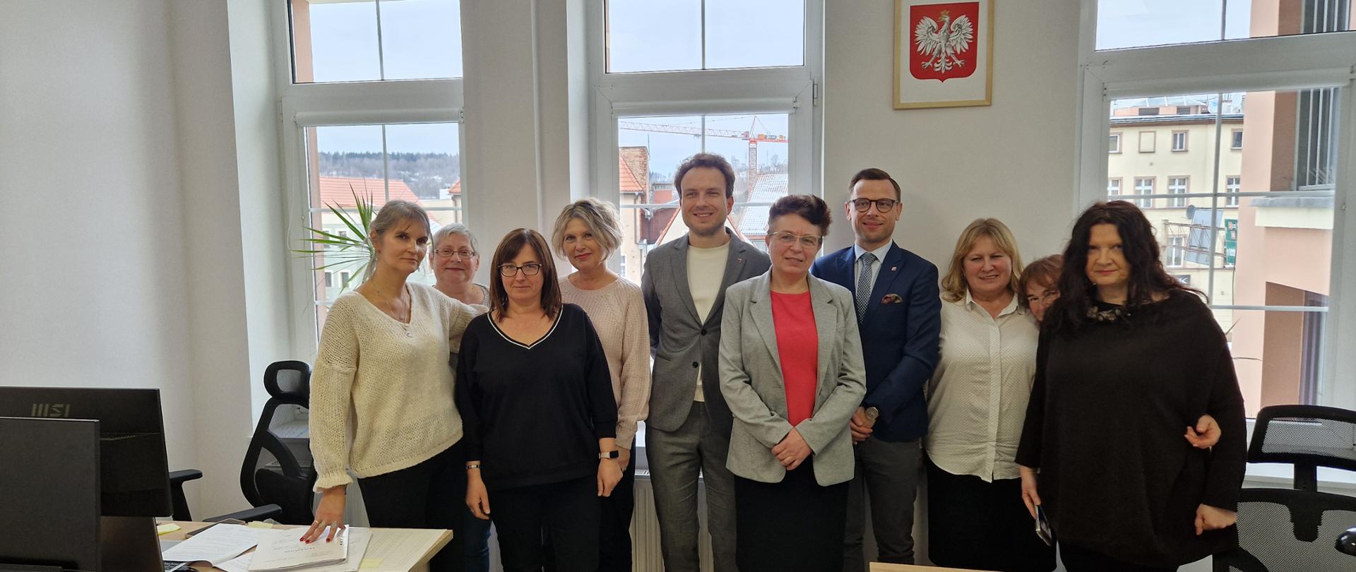 Grupa dziewięciu dorosłych osób stoi w sali biurowej przy biurkach. Osoby ustawione są w dwóch rzędach i patrzą w stronę aparatu. Pomieszczenie jest jasne, z dużymi oknami w tle, na ścianie widoczny jest herb Polski. Na stołach leżą dokumenty i drobne przedmioty biurowe. 