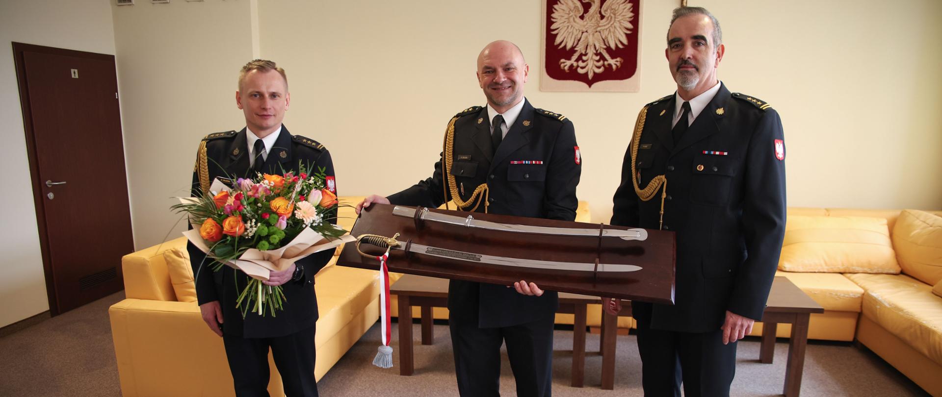 Żegnający się ze służbą starszy brygadier trzymający w dłoniach otrzymaną szablę oficerską wraz z pochwą umieszczoną na pamiątkowym tablo, Po jego lewej oraz prawej stronie stoją Zastępcy Komendanta Centralnej Szkoły PSP