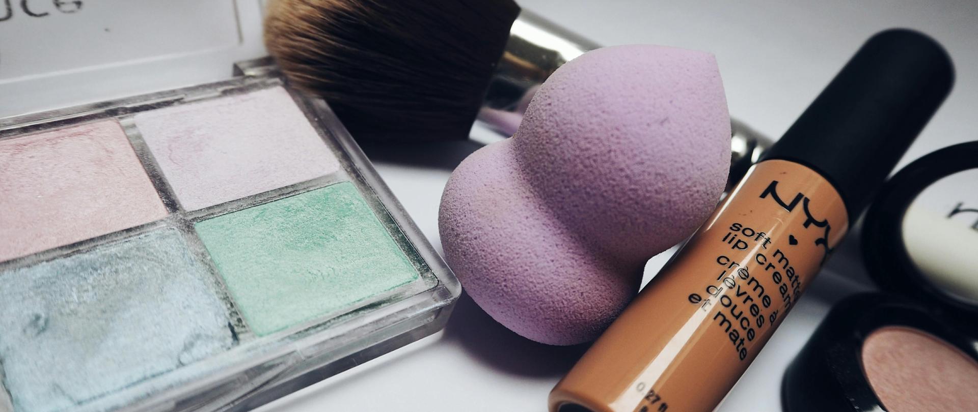 Z lewej strony pastelowe cienie do powiek, na środku różowa gąbka do makijażu oraz pędzelek do różu. Po prawej stronie róż do policzków oraz beżowy błyszczyk.