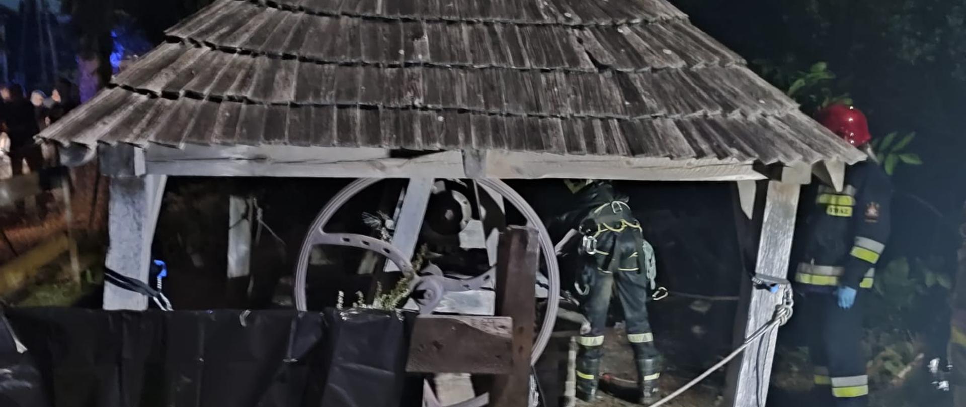 Zdjęcie przedstawia studnię zabezpieczoną drewnianym daszkiem z której ratownicy wydobyli poszkodowanego. Widać dwóch strażaków w czasie prowadzenia akcji.