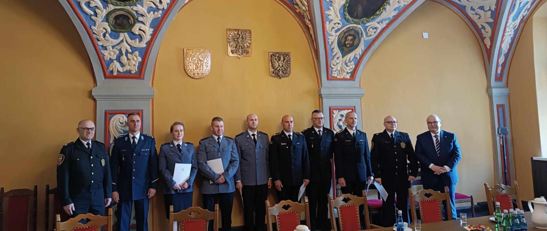 Zbiórka funkcjonariuszy w mundurach stojących w rzędzie w zabytkowym pomieszczeniu.