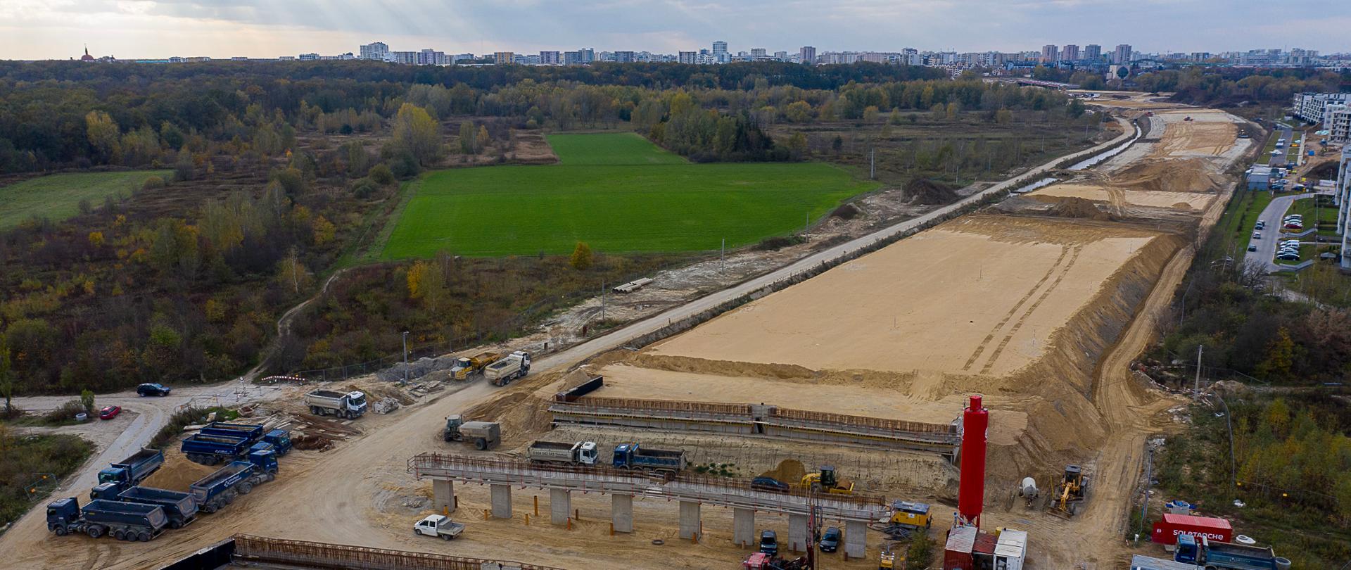 Zdjęcie przedstawia drogę ekspresową w budowie. Na zdjęciu widać rozległy plac budowy, na którym trwają prace nad nową drogą ekspresową. Widoczne są różne maszyny budowlane, takie jak koparki i ciężarówki, a także materiały budowlane. Droga jest jeszcze w fazie przygotowawczej, z wyraźnie zaznaczonymi pasami ziemi i piasku. W tle można dostrzec tereny zielone oraz zabudowania miejskie. 