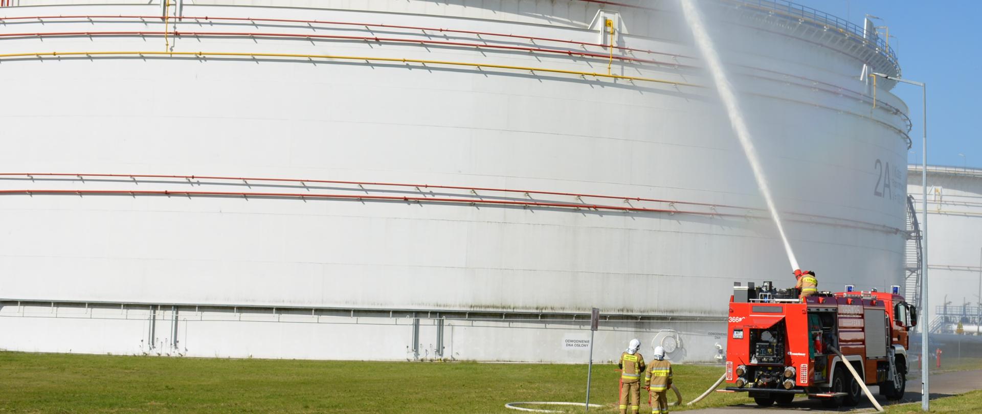 Z czerwonego wozu bojowego Państwowej Straży Pożarnej prowadzona jest pozorowana akcja gaśnicza: strumień wody skierowany jest na dach wysokiej na kilka metrów i pomalowanej na biało dużej konstrukcji. Obok wozu bojowego stoją dwaj funkcjonariusze Państwowej Straży Pożarnej.