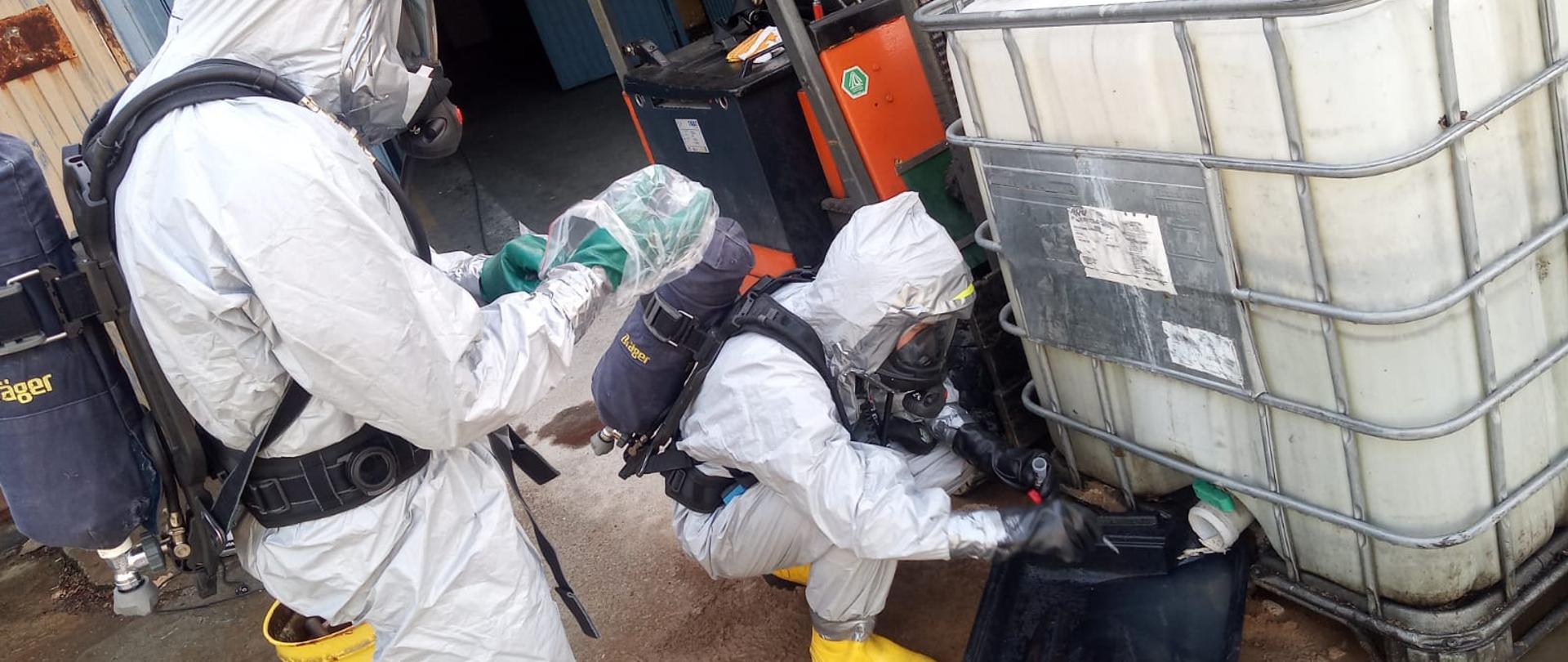 Strażacy w kombinezonach ochronnych pobierają próbki cieczy do analizy.