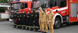 Strażacy stoją w pododdziale przed samochodami strażackimi na tle komendy