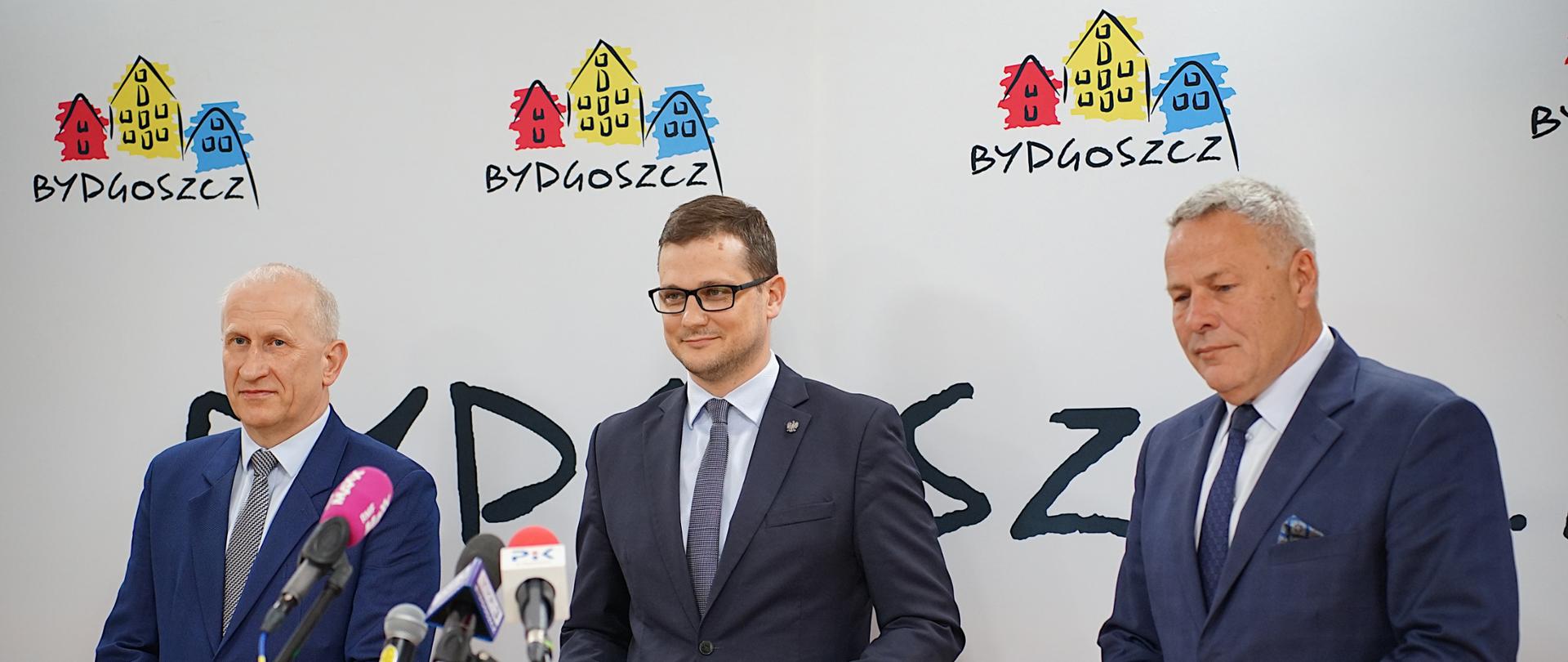 Na zdjęciu stoi trzech mężczyzn podczas konferencji prasowej. Wojewoda Kujawsko-Pomorski Michał Sztybel, Prezydent Bydgoszczy Rafał Bruski oraz przedstawiciel wykonawcy z bydgoskiej spółki Ebud-Przemysłówka. Mężczyźni stoją przed mikrofonami różnych stacji radiowych i telewizyjnych. W tle znajduje się oficjalna ścianka promocyjna z napisem „Bydgoszcz” oraz logo miasta.
Fot. B. Witkowski / UMB