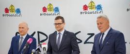 Na zdjęciu stoi trzech mężczyzn podczas konferencji prasowej. Wojewoda Kujawsko-Pomorski Michał Sztybel, Prezydent Bydgoszczy Rafał Bruski oraz przedstawiciel wykonawcy z bydgoskiej spółki Ebud-Przemysłówka. Mężczyźni stoją przed mikrofonami różnych stacji radiowych i telewizyjnych. W tle znajduje się oficjalna ścianka promocyjna z napisem „Bydgoszcz” oraz logo miasta.
Fot. B. Witkowski / UMB