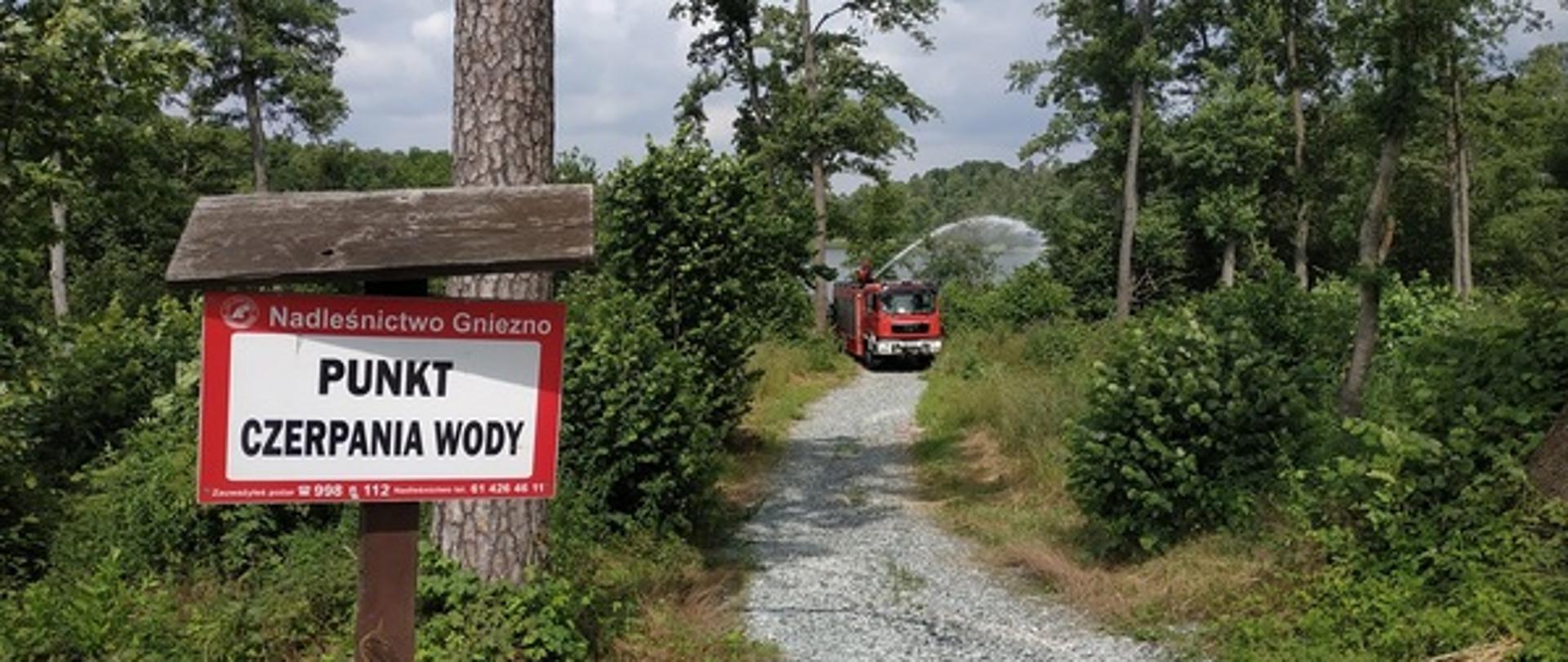 Tabliczka biała z czerwoną obwódką z czarnym napisem PUNKT CZERPANIA WODY zamontowana na drewnianym słupku. W tle kamienna droga i drzewa rosnące oraz czerwony wóz strażacki podający wodę z dachu pojazdu z działka wodnego.