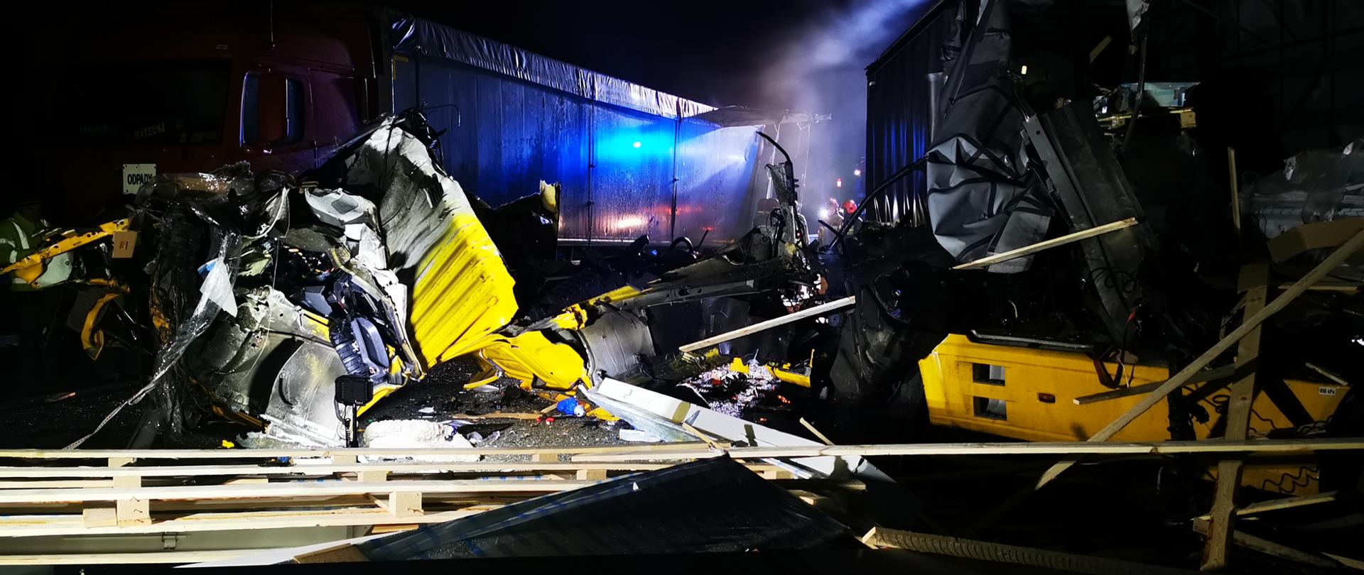 Zdjęcie przedstawia uszkodzoną kabinę samochodu ciężarowego. Z prawej strony widoczna uszkodzona naczepa wraz z przewożonym towarem rozrzuconym na drodze. W tle widać drugi samochód ciężarowy z uszkodzonym tyłem naczepy. 