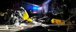 Zdjęcie przedstawia uszkodzoną kabinę samochodu ciężarowego. Z prawej strony widoczna uszkodzona naczepa wraz z przewożonym towarem rozrzuconym na drodze. W tle widać drugi samochód ciężarowy z uszkodzonym tyłem naczepy. 