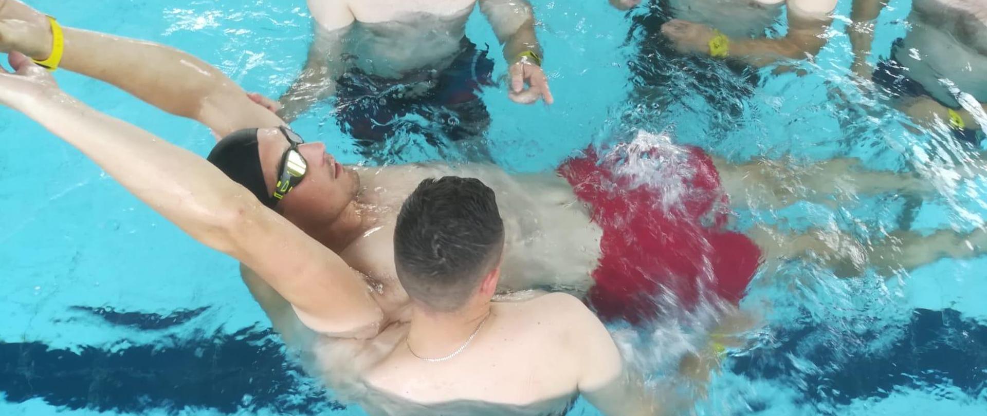 Jeden mężczyzna leży na wodzie w basenie a trzech stoi przy nim podczas zajęć szkoleniowych
