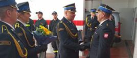 Komendant Powiatowy PSP w Rawiczu wręcza upominek nowo mianowanemu Dowódcy Jednostki Ratowniczo Gaśniczej. Po lewej stronie kolejni strażacy oczekują z bukietem kwiatów. W tle strażacy pełniący tego dnia służbę wraz z dowódcą uroczystości.