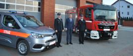 Dwóch funkcjonariuszy stoi przed budynkiem Państwowej Straży Pożarnej z jednej strony czerwony samochód marki MAN z drugiej strony srebrny samochód osobowy Toyota pomiędzy strażakami osoba w granatowym garniturze. Za nim bramy garażowe oraz budynek z cegły klinkier w oddali masz z flagą naraodową
