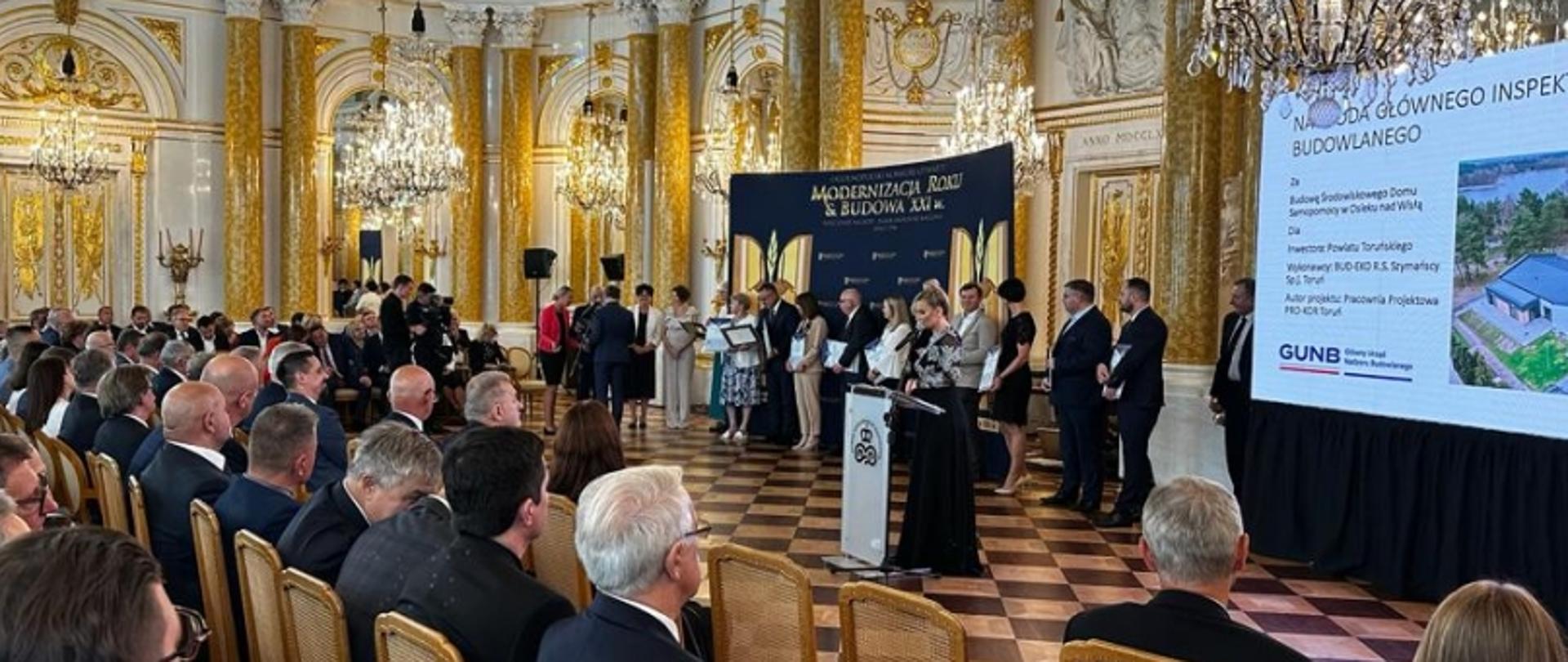 Sala z uroczystością wręczenia nagrody GUNB, ekran z informacjami o projekcie domu ekologicznego i uczestnikami siedzącymi naprzeciw sceny.