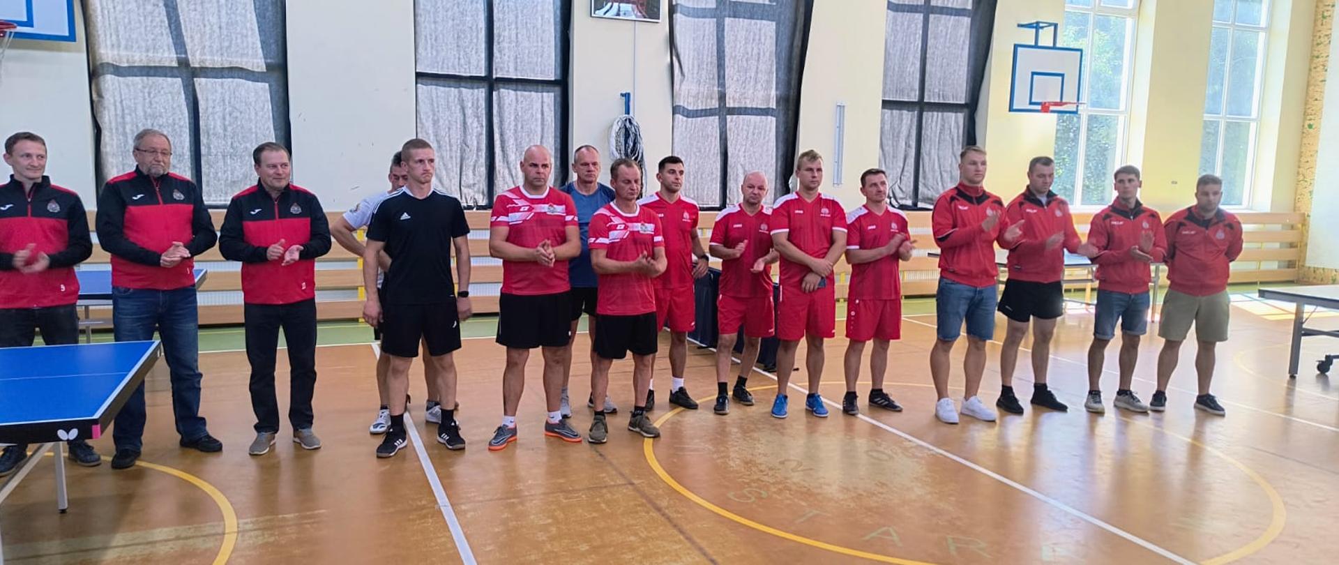 Kolorowa fotografia wykonana w pogodny dzień na sali gimnastycznej. Przedstawia strażaków w dresach sportowych i spodenkach znajdujących się na zbiórce po zakończeniu turnieju eliminacyjnego w tenisie stołowym.
