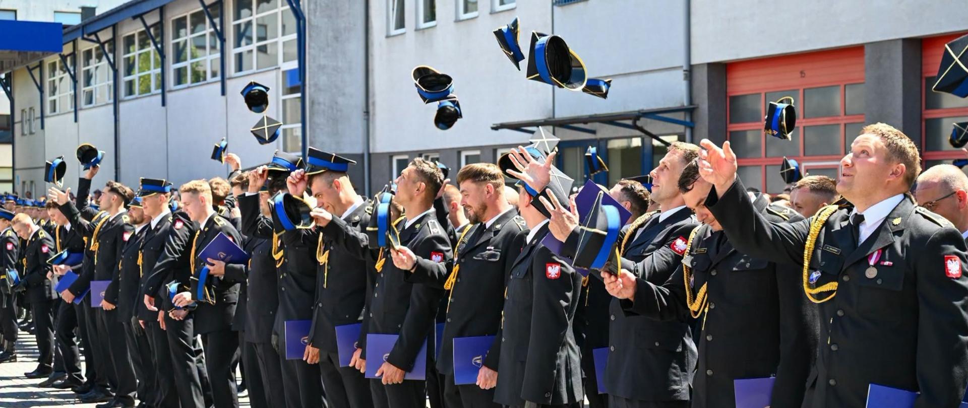 Grupa strażaków Państwowej Straży Pożarnej w galowych mundurach stoi w szeregu na placu przed budynkiem komendy. Wszyscy z uśmiechem i radością podrzucają w górę swoje czapki rogatywki podczas uroczystego apelu. W rękach trzymają niebieskie foldery lub dyplomy. W prawym dolnym rogu widnieje podpis: „st. str. kdt Piotr Krzak | Państwowa Straż Pożarna”.