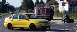 ZDERZENIE SKUTERA Z OSOBÓWKĄ. Na zdjęciu widzimy zderzenie samochodu osobowego ze skuterem. Żółty samochód ma rozbity przód, na jezdni leży rozbity skuter, w tle stoi samochód strażacki.