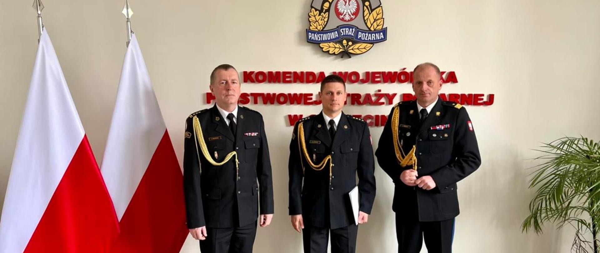Trzech funkcjonariuszy na tle ściany z napisem "Komenda Wojewódzka PSP w Szczecinie", z lewej strony dwie flagi Polski, z prawej strony kwiat.
Od lewej, st. bryg. Krzysztof Makowski, nadbryg. Jarosław Tomczyk, st. kpt. Krzysztofow Azierski.