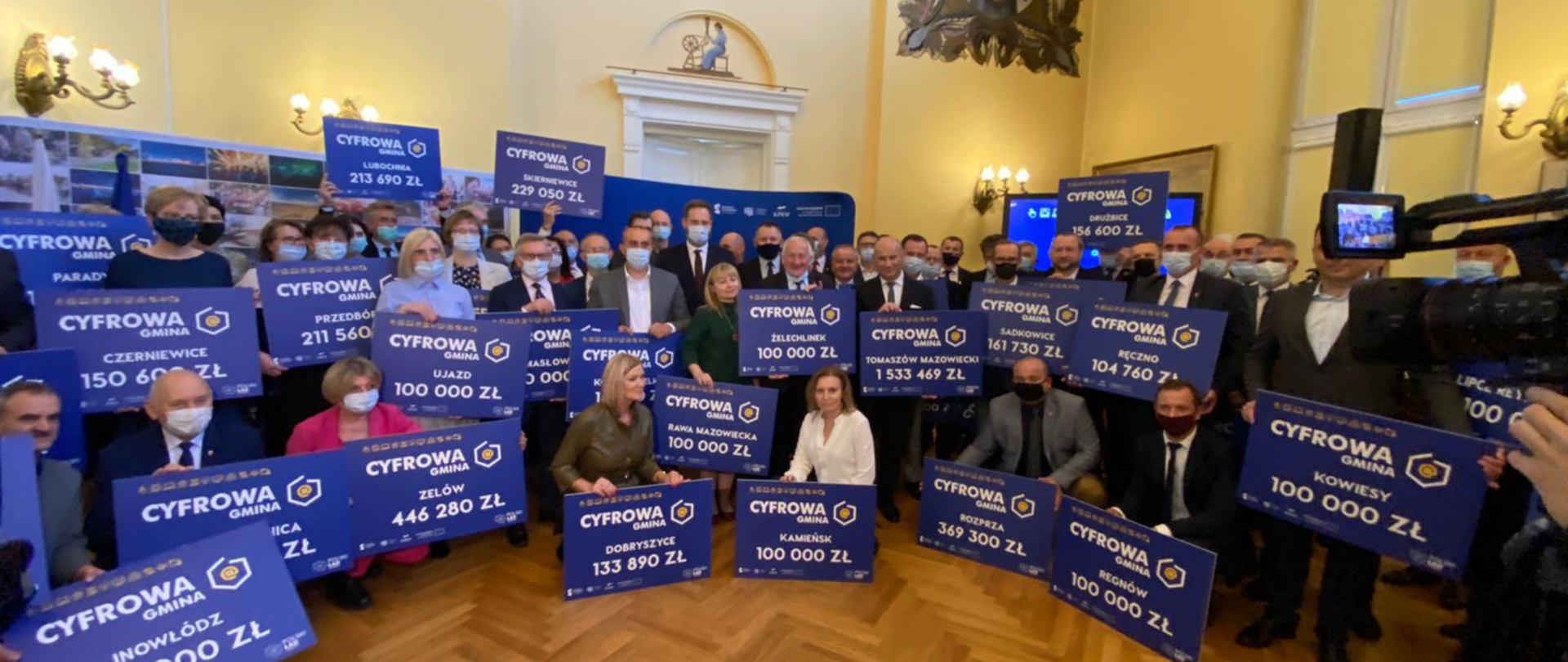 Minister Janusz Cieszyński z samorządowcami z woj. łódzkiego. Samorządowcy trzymają symboliczne czeki.