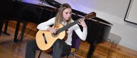 Na zdjęciu znajduje się uczennica grająca na gitarze, w tle stoi czarny fortepian na drewnianej podłodze - scenie, ściana koloru jasnego
