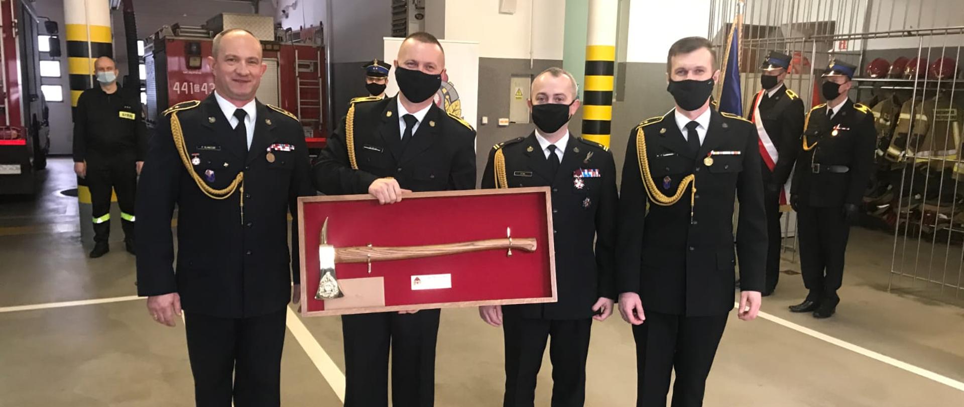 Zdjęcie st. ogn. Mariusza Dejk trzymającego pamiątkowy topór, w obecności komendanta powiatowego PSP w Kartuzach oraz dowódcy i zastępcy dowódcy zmiany I JRG Kartuzach. 