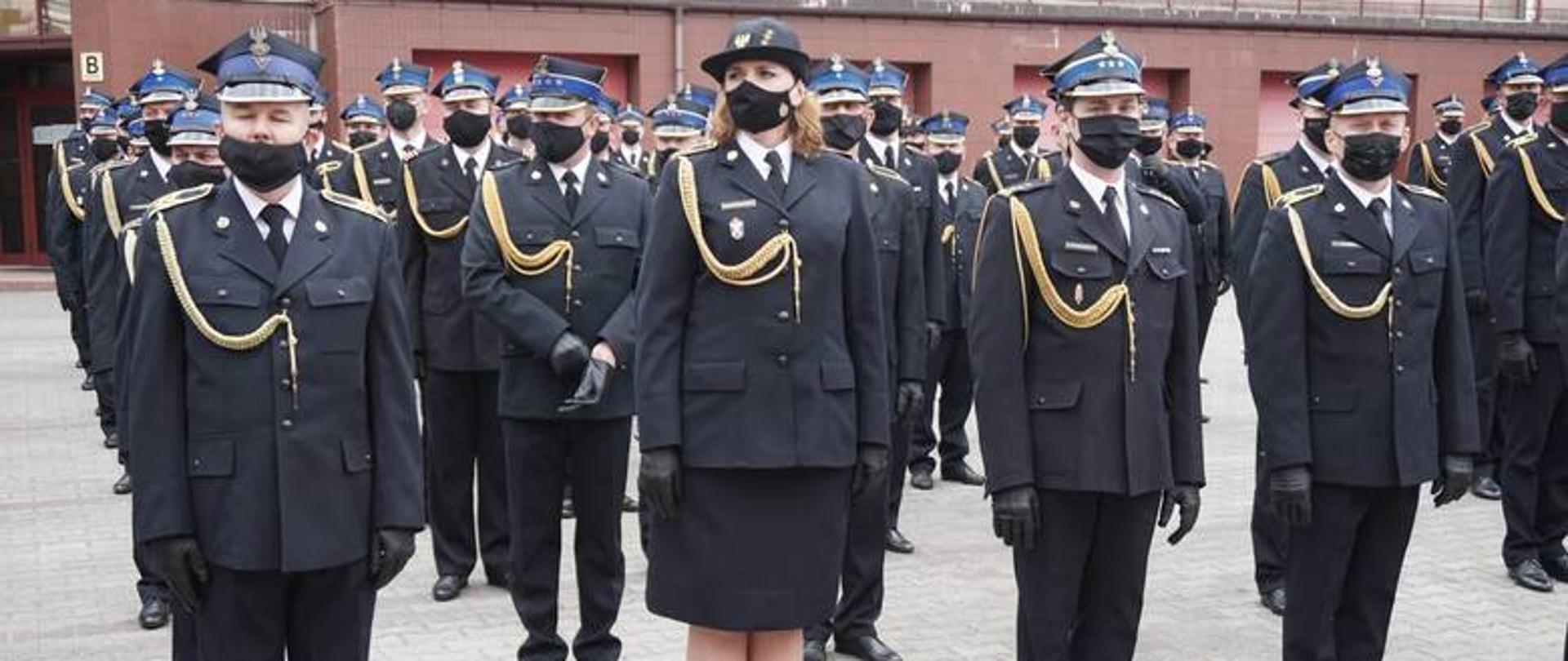 Zdjęcie przedstawia strażaków Państwowej Straży Pożarnych w umundurowaniu wyjściowym ze sznurem galowym stojących na zbiórce na placu wewnętrznym Komendy Wojewódzkiej Państwowej Straży Pożarnej w Łodzi. Każdy strażak posiada dodatkowo czarną maseczkę zakrywającą twarz i nos oraz rękawice również koloru czarnego.